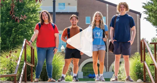 Four students standing outdoors in front of the SAU Library, smiling.