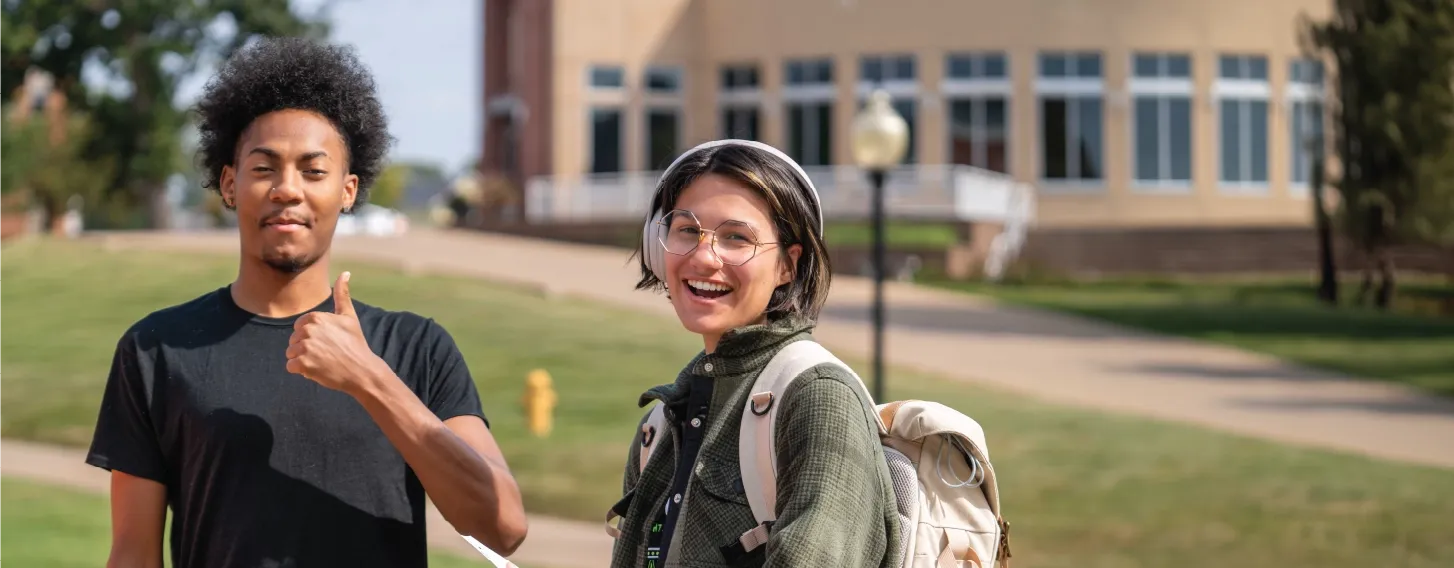 Two students standing outdoors, smiling, with one giving a thumbs up sign.